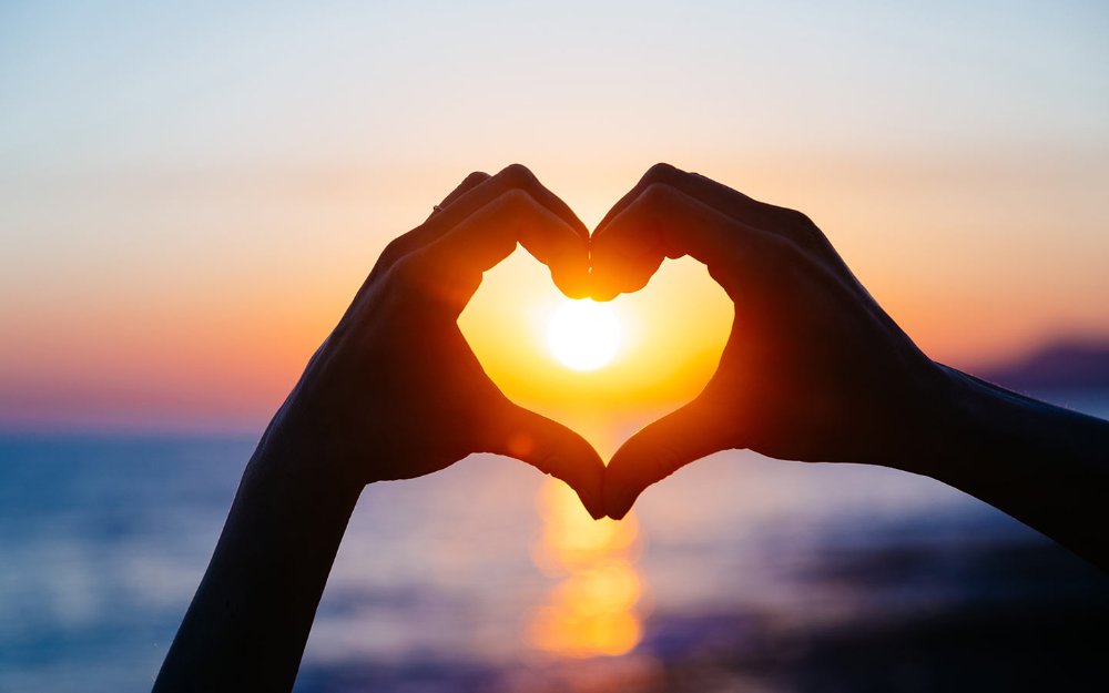 Hände, die vor dem Hintergrund eines Sonnenuntergangs ein Herz formen, symbolisieren Romantik und Liebe.